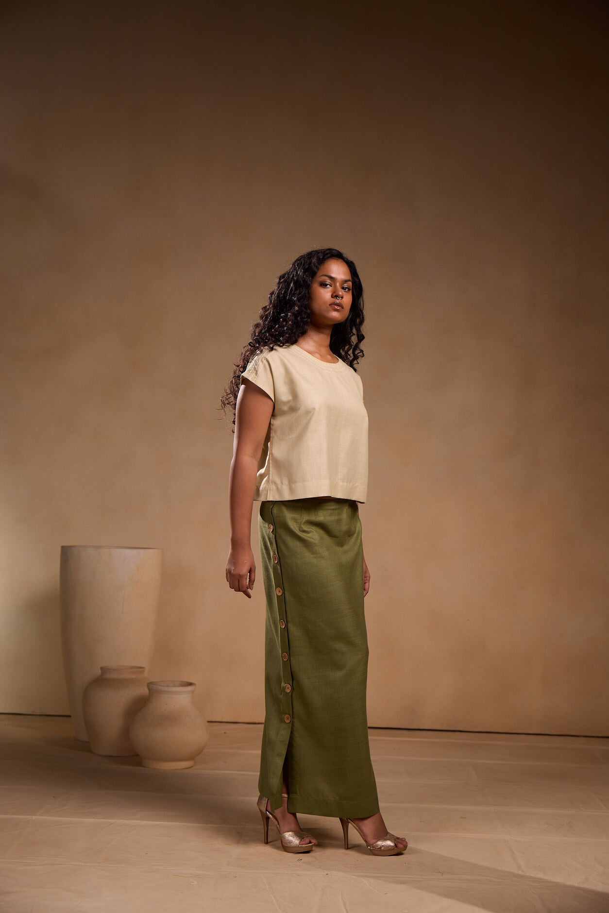 Woman wearing a beige top and green skirt standing against a beige wall with decorative vases.