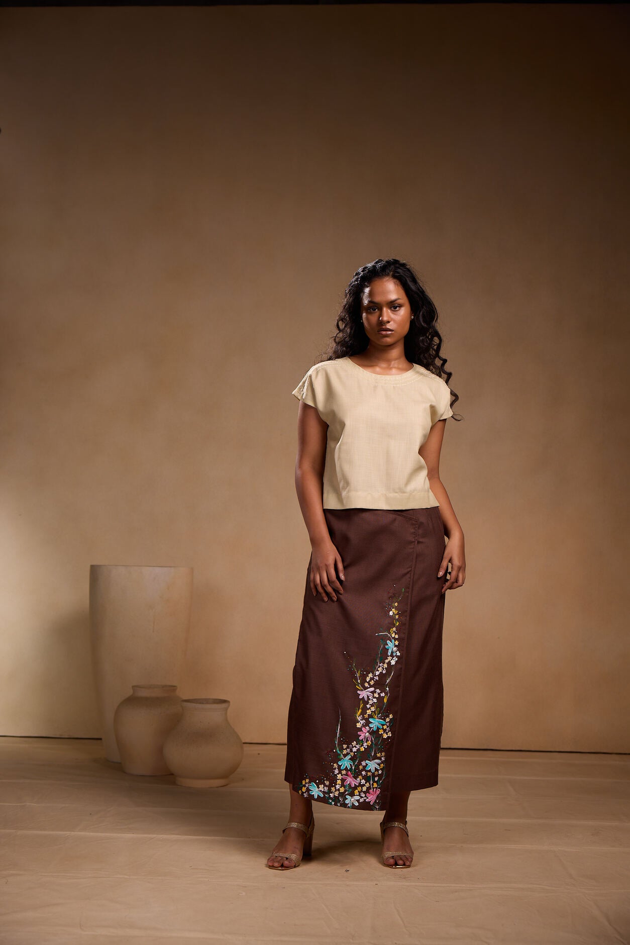Woman wearing a beige top and brown skirt with floral embroidery against a beige background